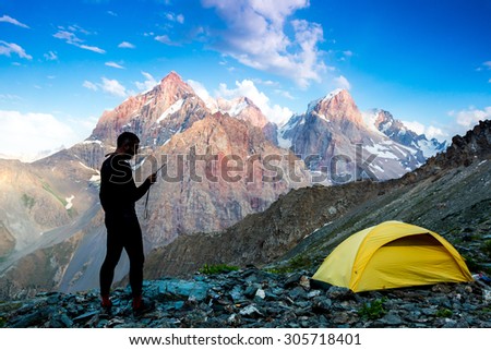 Explorer talking via radio station.\
Silhouette of man in wild mountain landscape walk along yellow camping tent holding radio transmitter connection with team blue sky sunny evening