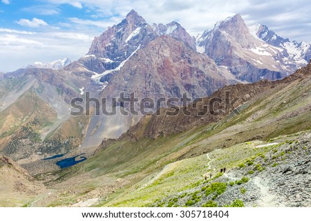 White mountain footpath and group of hikers.\
Mountain landscape with green grass and red orange rocky peaks white dusty trail and group of travelers walking toward camping tents
