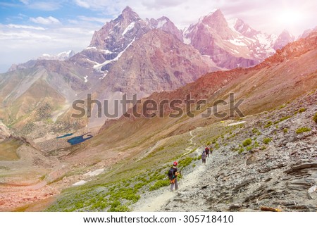 White mountain footpath and group of hikers.\
Mountain landscape with green grass and red orange rocky peaks white dusty trail and group of travelers walking shining sun background