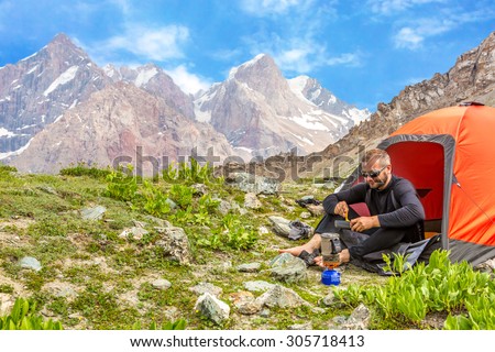 Traveling man eating meal.\
Hiker sitting in his orange camping tent and having lunch stove and cooking gear mountain landscape on background