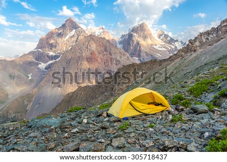 Yellow tent on mountain landscape.\
Small camping single tent located on rocky terrain stone surface and high mountain hills and peaks on background