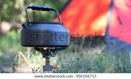 Campfire. \
Close-up image of burning gas stove and kettle with boiling water located on green grass lawn outdoor red orange tent on background focus on stove and fire tent is blurred