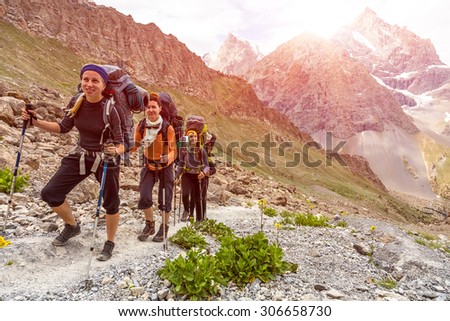 Group of three hikers on trail.\
Mountain landscape and people walking with poles backpacks and other gear along dusty Asian trail with green grass and orange rocks around