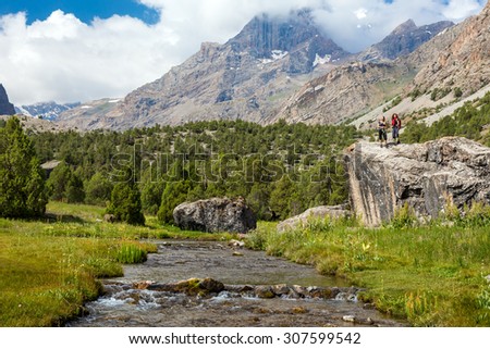 Mountain Stream and Female Hikers on Rock.\
Two People Staying on Rock above Flow of Creek with green Meadows and Forest Mountain View massive Clouds Blue Sky on Background