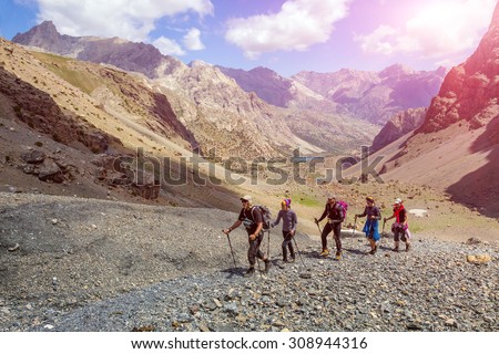 Diverse group of hikers in mountain terrain.\
People walking along rocky footpath positive faces trekking gear poles sporty clothing bright panorama blue sky sunny day background