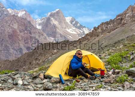Cool man eating lunch in mountain hike.\
Climber sitting in yellow single tent set up on rocky terrain with camping stove plate bread mug eat cooked lunch high mountains and blue sky background