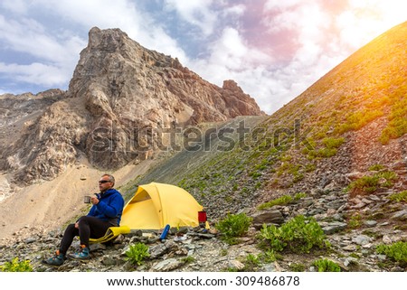 Lunch of alpine climber.\
Hiker sitting aside yellow camping tent and having lunch stove and cooking gear mountain landscape shining sun sunbeams on background