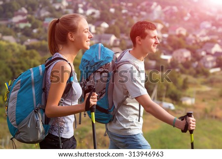 Excited Travelers Young Man and Woman Traveling Outdoor Expressing Fun and Pleasure with Backpacks Walking Poles Sticks and Casual Sporty Style Clothing Suburban Park Landscape Shining Sun Background