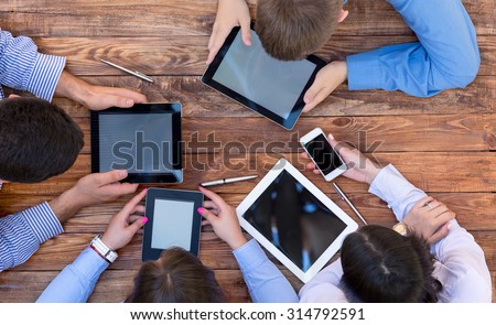 Men and women Staring Intensively on Screens of Digital Gadgets.\
Natural Rough Wooden Plank Desk and Four People Holding Working Electronic Devices Tablet PC Book Notepad Smart Telephone
