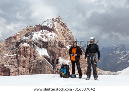 Two Climbers on Snow Pass\
Tall and Short Men with Climbing Gear and Expedition Clothing Backpacks on Glacier with High Steep Rock Peak and Stormy Sky on Background