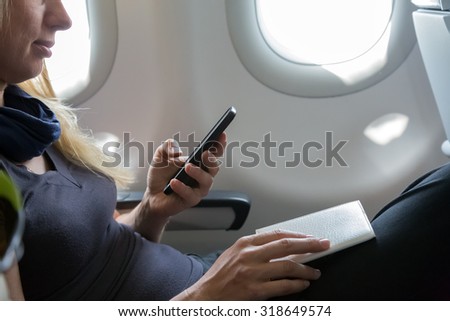 Air Flight Passenger Sitting in Aircraft\
Casual Clothing Woman in Plane Seat Browsing Her Smart Phone and Holding Documents for Immigration Formalities Airplane Windows on Background