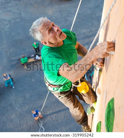 Aged Person Practicing Extreme Sport\
Elderly Male Climber Makes Hard Move Looking High Up on Outdoor Climbing Wall Sport Competitions Very Emotional Face Belaying Partner Fans Staying on Remote Ground