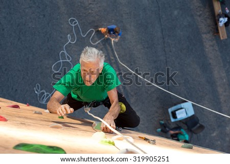 Mature Athlete Trying to Reach Out Next Climbing Hold\
Aged Climber Makes Hard Move on Outdoor Vertical Gym Sporty Clothing Intense Emotional Face Belaying Partner on Background