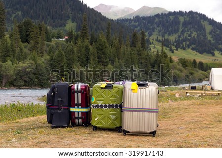 Travel Bags Staying on Wild Grassy Meadow \
Mountain View with Forest Hills and River Stream and Many Suitcases of different type and colors