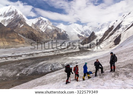 Group of Hikers Walking on Snow and Ice Terrain.\
Large Group of People Sport Clothing Climbing Gear High Altitude Boots Going Up Mountain Peaks Sunlight Cloud Sky Massive Glacier Flow on Background