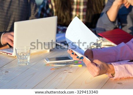 Hands of Businesswoman Holding Paper Prints of Presentation and Discussing with Partners Sitting around Wood Table with Laptop Pencils Telephone Glass of Water and Notepads