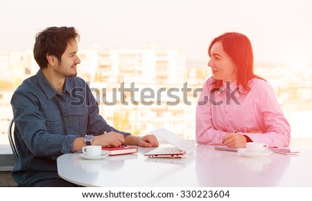 Man and Woman Casual Clothing Sitting at White Round Table with Coffee Mugs Laptop Notepads and Telephone Discussing Smiling Making Hand Notes Urban Landscape on Background Shining Sun Back-light