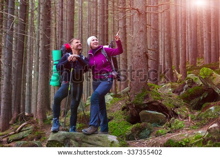 Man and Woman Hikers Staying on Stone Covered by Moss in Dense Old Forest Smiling and Pointing with Backpacks and Trekking Poles Sunbeams Shining throw Trees