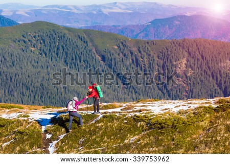 Two Hikers in Mountain Landscape Man helping Woman to climb on steep Ridge Forest Hills and shining Sun on Background