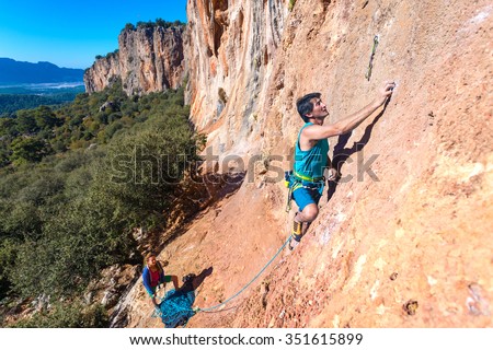 Team of Climbers Man and Woman ascending orange bright rocky Wall with rope and gear Male Leading Female belaying blue Sky and green Forest