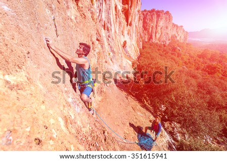 Team of Climbers Man and Woman ascending orange bright rocky Wall with rope and gear Male Leading Female belaying blue Sky and green Forest