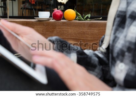 Hands of Person browsing tablet PC screen close up with Coffee Mug and Pomegranate and Orange on foreground, focus on fruits and white coffee Cup