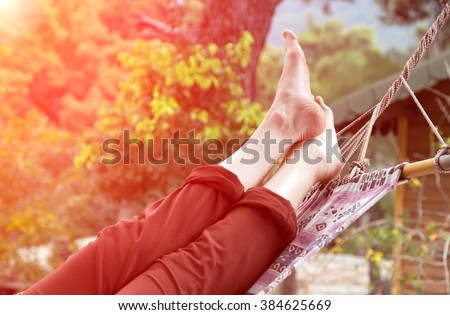 Person relaxing lying in Hammock at rural cottage garden female legs green flora and wooden hut on background