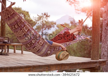 Person relaxing lying in Hammock at rural cottage garden female legs green flora and wooden hut on background
