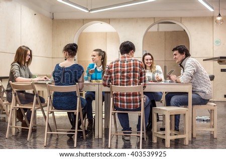 Many Students in casual Clothes Men and Women sitting at wooden Table inside Campus Chat Room area talking laughing working