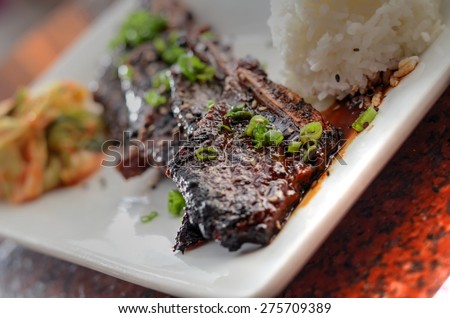 Korean short ribs on a rectangular plate with rice and kimchi in the background