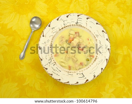 A serving of chicken dumpling soup with vegetables in a bowl with spoon on a yellow floral print tablecloth.
