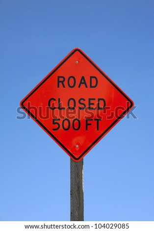 A bright orange road closed five hundred feet sign on a wood post against a blue sky.