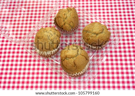 A group of four fresh apple spice muffins in the plastic container on a red and white checkerboard cloth.