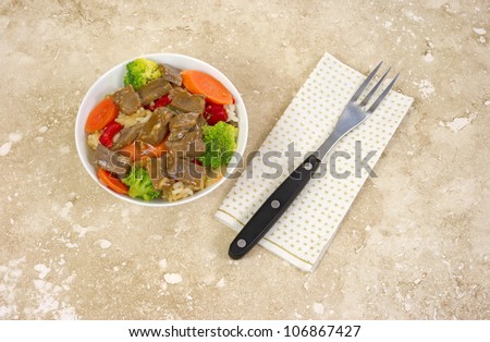 A small bowl of beef teriyaki with vegetables and a fork on paper napkin atop a marble tabletop.