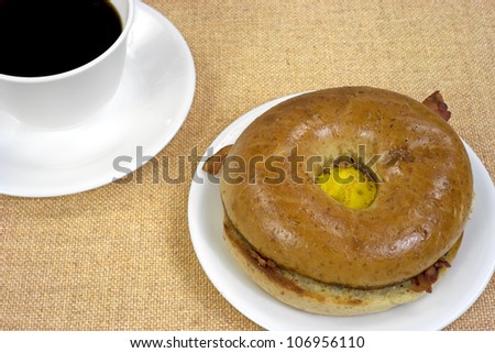 A whole wheat bagel with bacon eggs and cheese on a white plate with a cup of black coffee to the side on a tan cloth background.