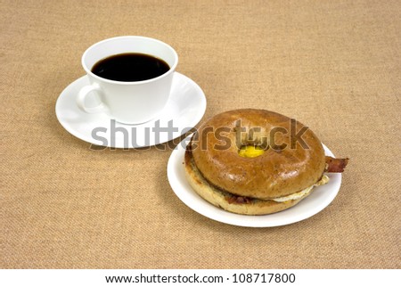 A cup of black coffee with a freshly made wheat bagel filled with bacon egg and cheese on white plate and saucer.