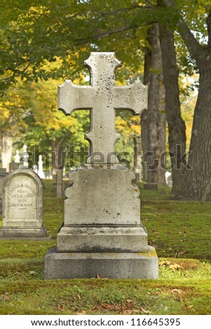 The back of a cross shaped tombstone in a cemetery with trees and grass as fall is just beginning.