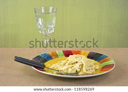 A serving of Fettuccine Alfredo TV dinner on a bright plate with black handle fork and a goblet of water in the background.