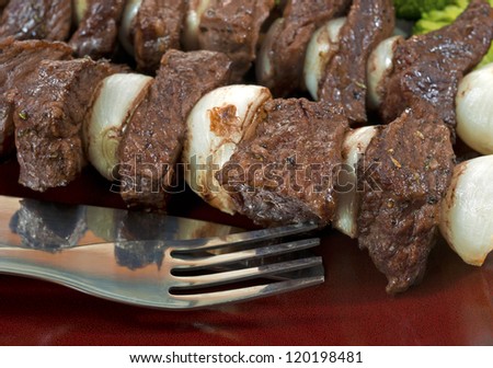 Close view of a meal of cooked beef and onion kabobs on a red plate with fork and knife.