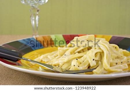 Close view of a TV dinner meal of Fettuccine Alfredo on plate with fork with a crystal goblet in the background.