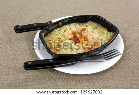 A TV dinner of shrimp and angel hair pasta with vegetables in a black microwave dish on a white plate with fork and knife.