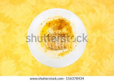 A small portion of haddock that has been microwaved too long on a dish atop a yellow floral tablecloth background.