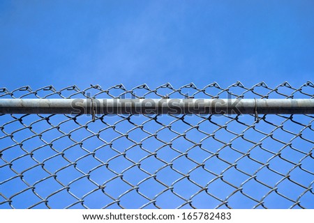 A chain link fence with a sturdy bar and fastenings against a blue sky with wispy clouds.