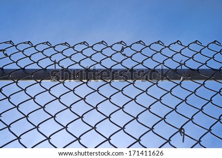 A chain link fence with a sturdy bar behind the mesh and fastenings against a blue sky with wispy clouds.