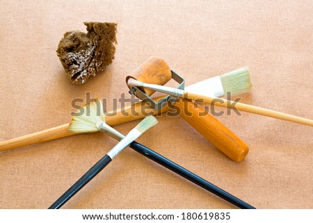 A group of artist brushes, a sponge and roller on a tan burlap background.