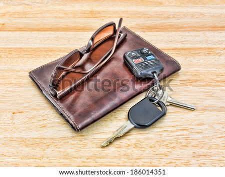 A set of car keys and tinted sunglasses atop a brown leather wallet on wood tabletop.
