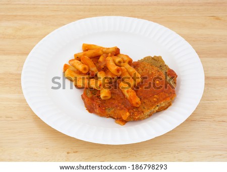 A cooked TV dinner of breaded chicken with tomato sauce and pasta on a paper plate atop a wood tabletop.