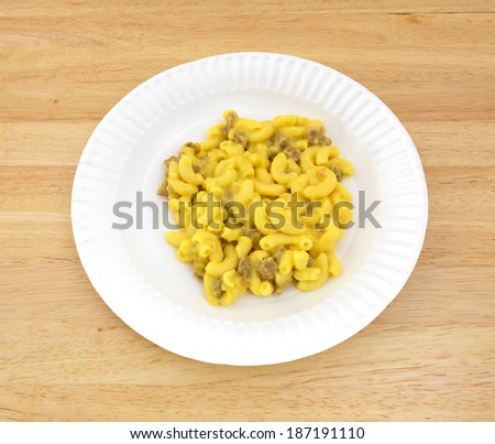 A paper plate with a serving of macaroni and cheese with meat on a wood dining tabletop.