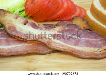 Close view of cooked bacon with lettuce tomatoes and white bread on a wood cutting board.