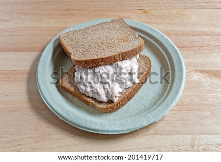 A freshly made tuna sandwich on whole wheat bread on a paper plate atop a wood table.